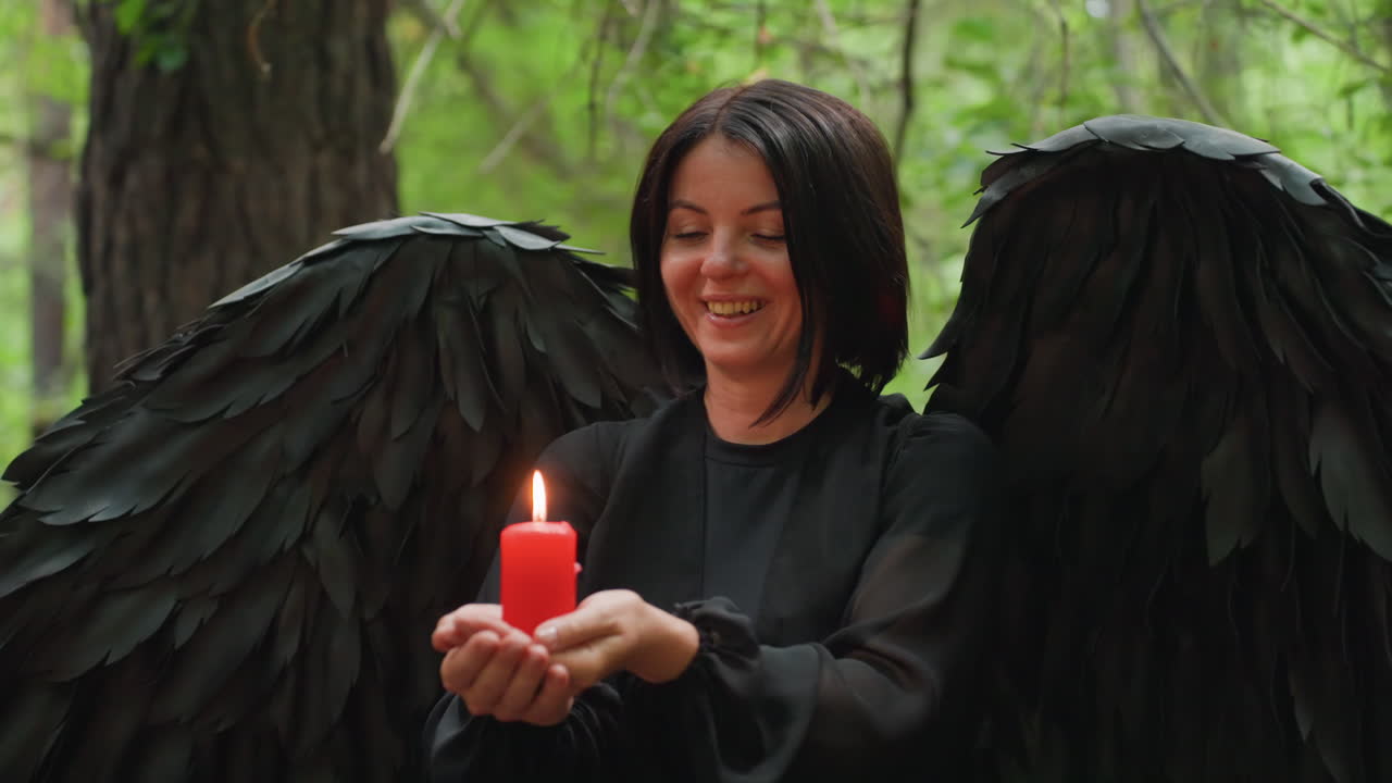Lady with black wings holds burning red candle in hands while standing in quiet forest, performing gentle spiritual ritual surrounded by soft light, tranquil atmosphere of devotion and faith