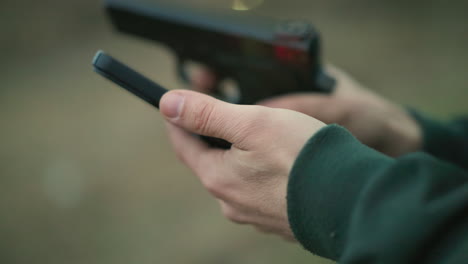 a close-up view of a man in a green jacket holding a cell phone, operating it and a gun, focusing on his hands