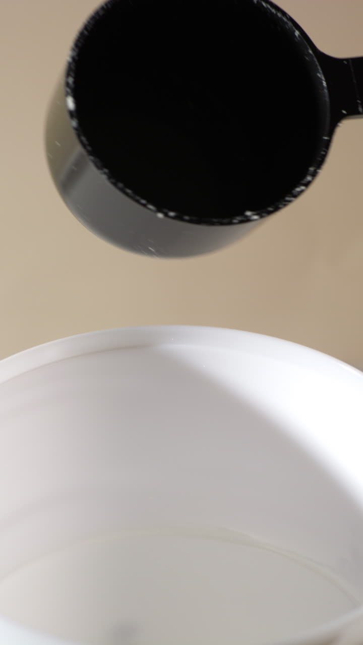 A scoop of vanilla or banana protein powder is shown mid-pour into a white cup, with fine powder cascading downward in soft natural light against a clean, minimal background for a health-focused look.