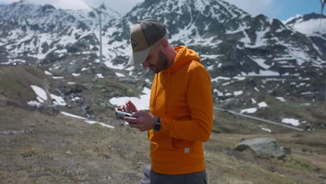 caucasian bearded man standing while holding and setting parameters on drone remote controller with snow-capped mountains and wind turbine on background