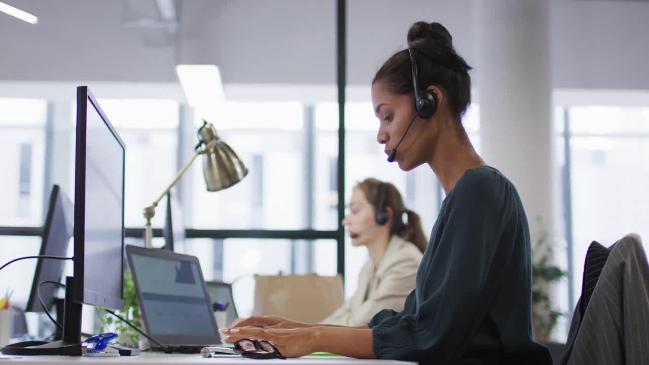 Geschäftsfrau gemischter Herkunft sitzt am Schreibtisch, telefoniert mit einem Headset und nutzt einen Computer in einem geschäftigen Büro