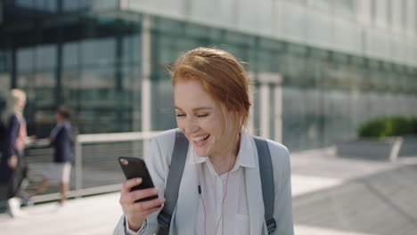 Retrato-De-Una-Alegre-Mujer-De-Negocios-Pelirroja-En-Prácticas-Usando-Auriculares-Escuchando-Música-Enviando-Mensajes-De-Texto-Navegando-Usando-La-Aplicación-De-Redes-Sociales-Para-Teléfonos-Inteligentes-Riéndose-Leyendo-Mensajes