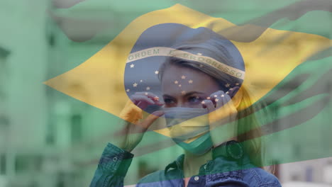 animation of flag of brazil waving over caucasian woman wearing face mask in city street