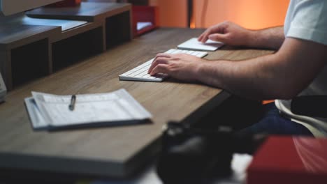 creative person sitting at his home office modern wood desk typing on a white wireless computer keyboard, clicking a mouse trackpad and writing down notes on notepad with an orange light in background