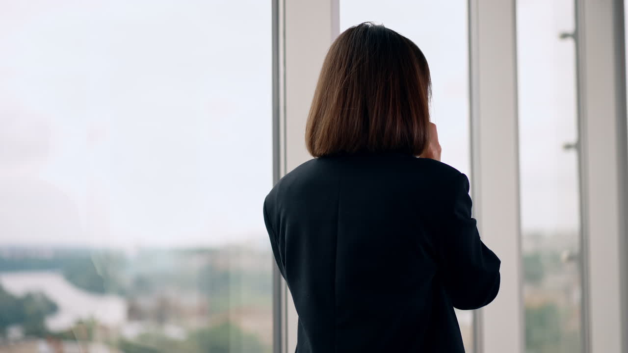 Brunette business lady having phone conversation in the office. Woman looks at the window, speaks on the smartphone and laughs.