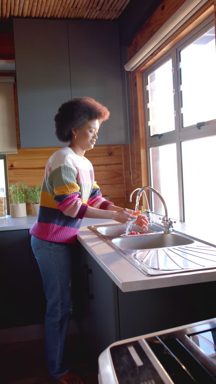 Video verticale di una felice donna afroamericana che sciacqua le verdure in una cucina soleggiata, al rallentatore.