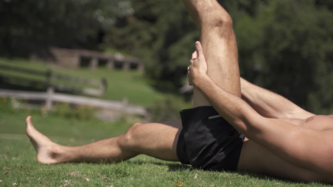 4K Young caucasian fit attractive male exersicing outdoors with green vegetation background hamstrings mobility exercise