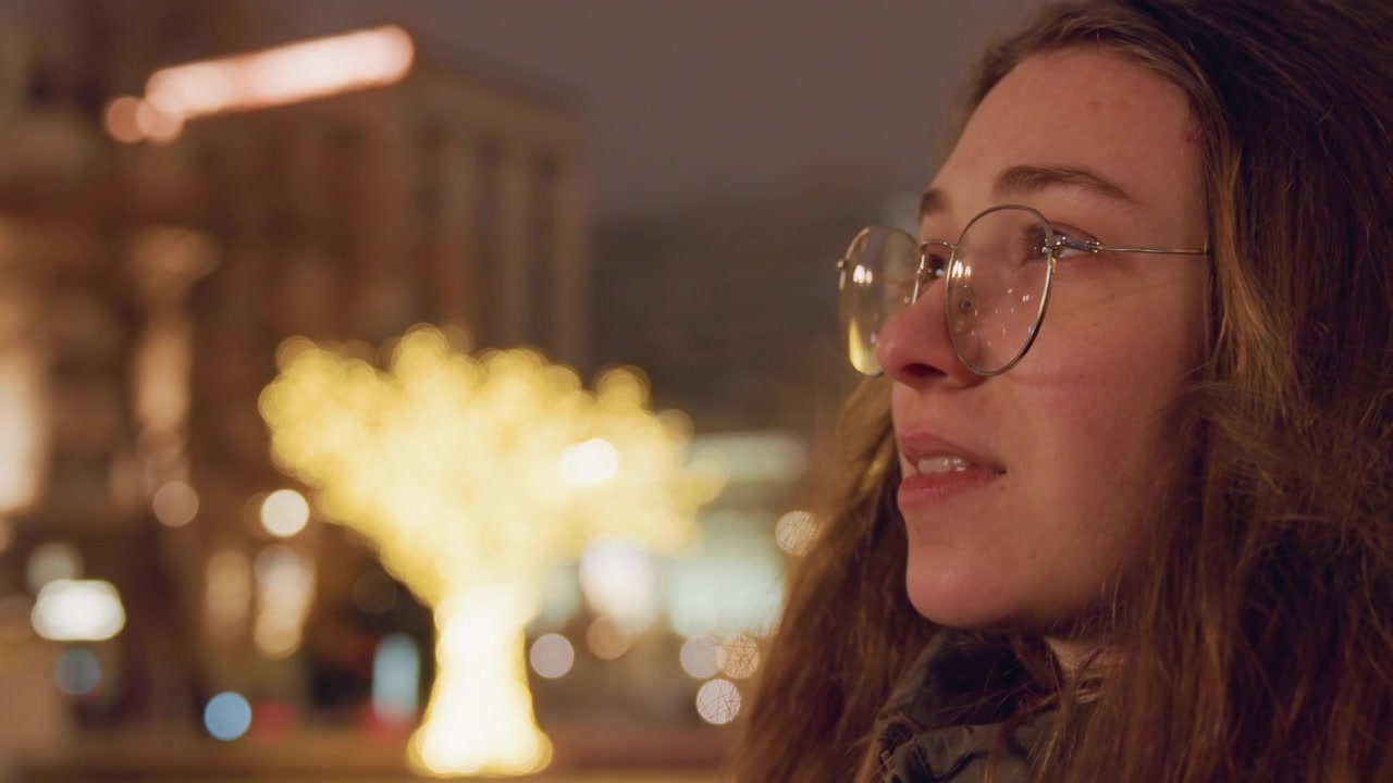 Profilo di donna caucasica con occhiali sotto un bokeh dorato di alberi, calda notte cittadina, espressione ravvicinata di tranquilla meraviglia, guance rosee, capelli fluenti, skyline urbano e luci dei lampioni sfocati, atmosfera artistica
