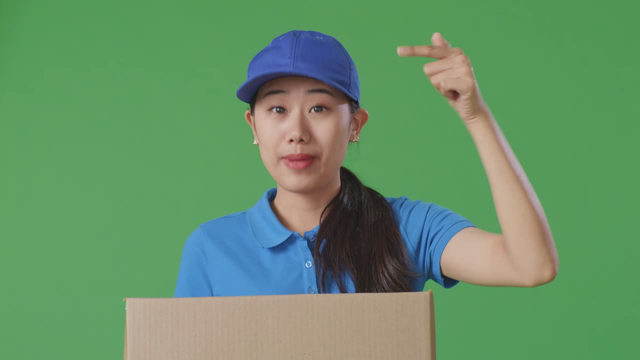 푸른 유니폼을 입은 아시아 여성 택배사가 스튜디오에서 녹색 스크린 배경에 상자를 배달하는 동안 카메라에 미소 짓고 이야기합니다.