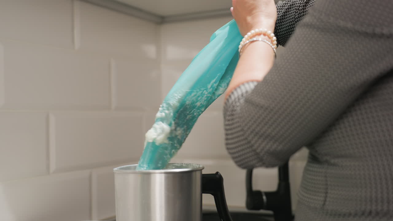 Creative lady presses creamy flakes from blue piping bag into metal bowl on kitchen counter while preparing ingredients for recipe, wearing bracelet and focused on culinary task with precision