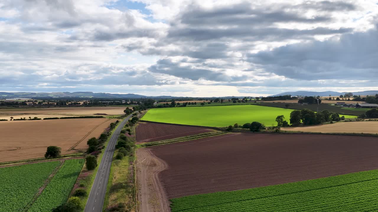 Drone footage glides above patchwork fields and a rural road in Kinross, Scotland, under partly cloudy skies with soft, natural daylight