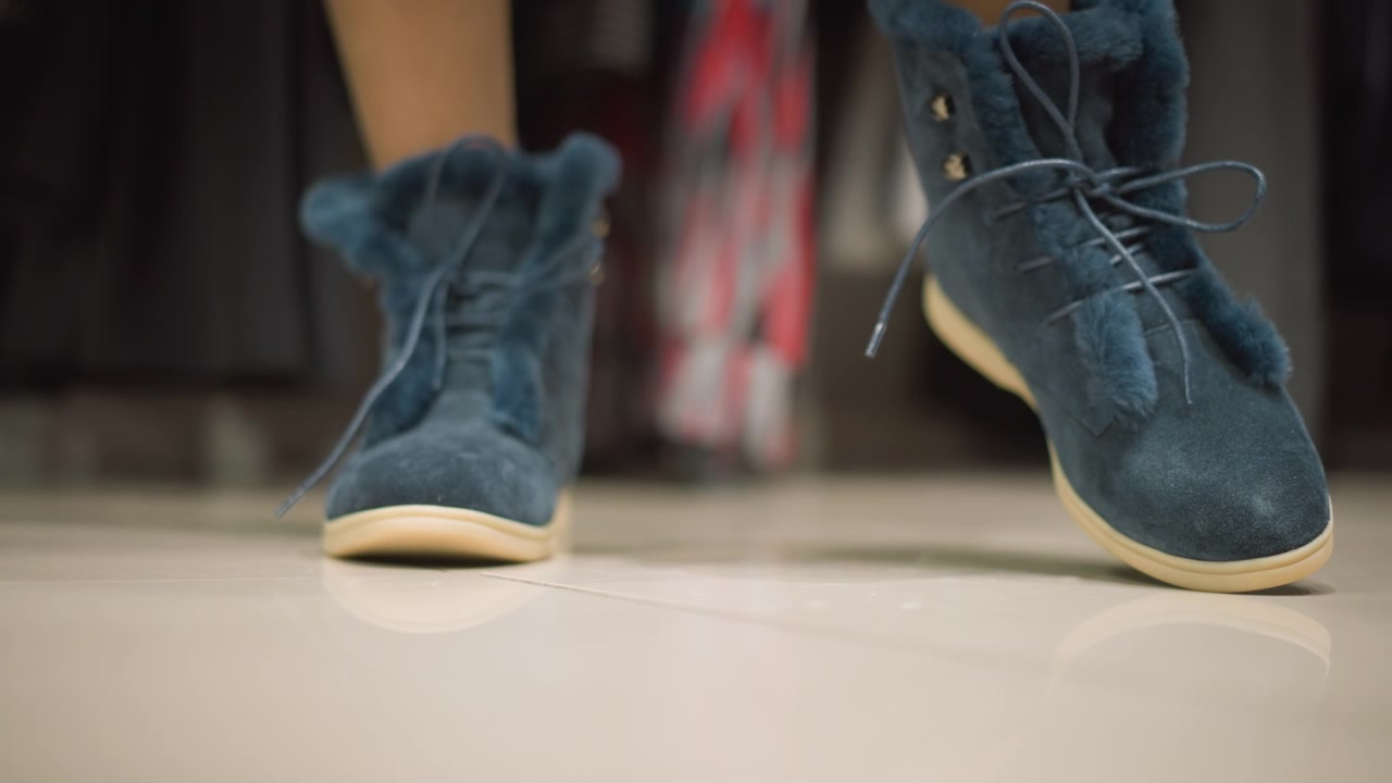 Close-up de botas de tornozelo de camurça azul com acabamento de pele e cadarços em piso de cor clara. A câmera foca nos detalhes da textura, mostrando leve movimento enquanto os pés mudam de posição em ambiente interno. Iluminação suave.