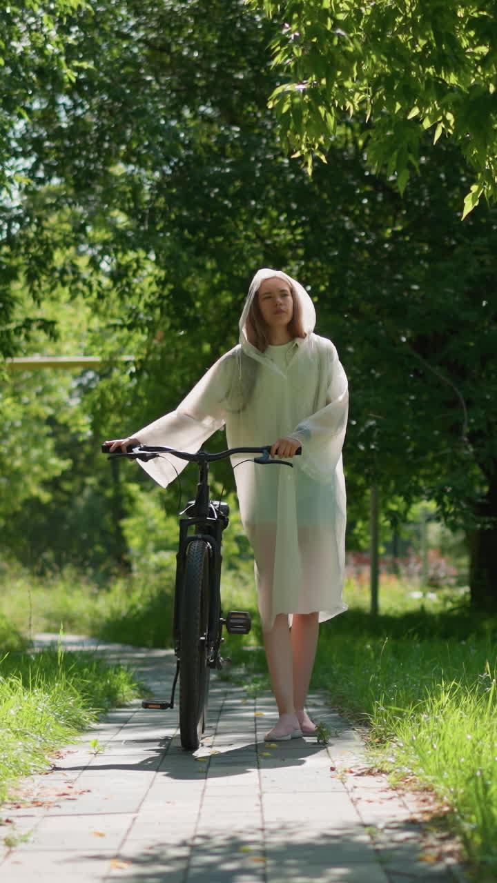 반투명한 레인코트를 입은 젊은 여성은 초록색으로 둘러싸인 빛으로 빛나는 길을 따라 자전거를 타고 산책합니다.