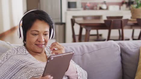 Mujer-Mayor-Afroamericana-Usando-Audífonos-Usando-Una-Tableta-Digital-Sentada-En-El-Sofá-En-Casa