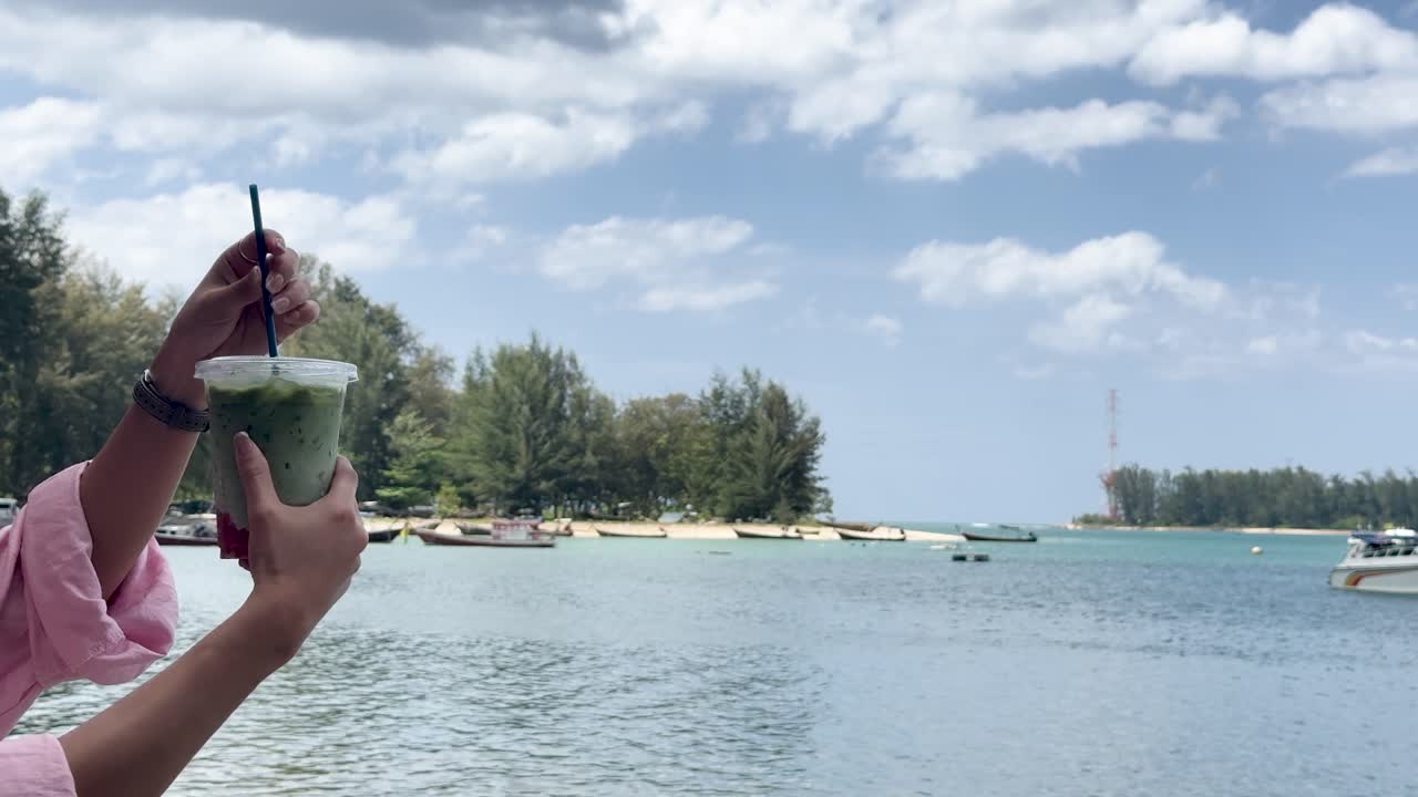 Person stirs green iced beverage with straw, tropical beach and blue sky in background