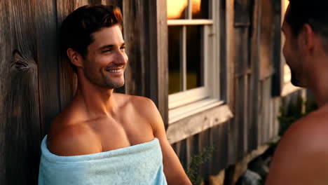 man in a towel smiling outdoors next to a rustic cabin at sunset