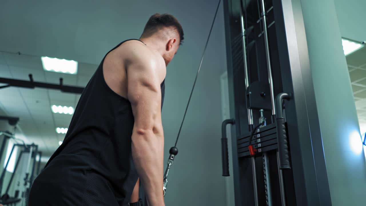 Muscular man trains on the simulator in fitness center. Strong athlete pull bar with rope and weights on fitness machine indoors.