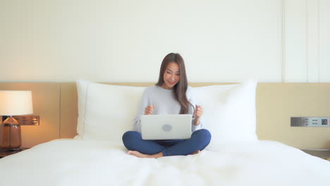 Sitting-crossed-legged-on-a-huge-hotel-room-bed-and-working-diligently-on-her-laptop-a-pretty-young-woman-reacting-to-her-successes