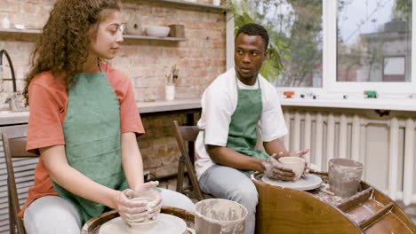 employees wearing green apron modeling ceramic pieces on potter wheel in a workshop while talking to each other 3