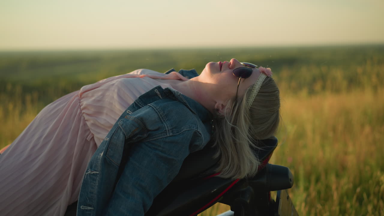 A woman lies back on her bike wearing sunglasses, adjusting her denim jacket as she relaxes in an open grassy field under the sun, enjoying the peaceful nature around her