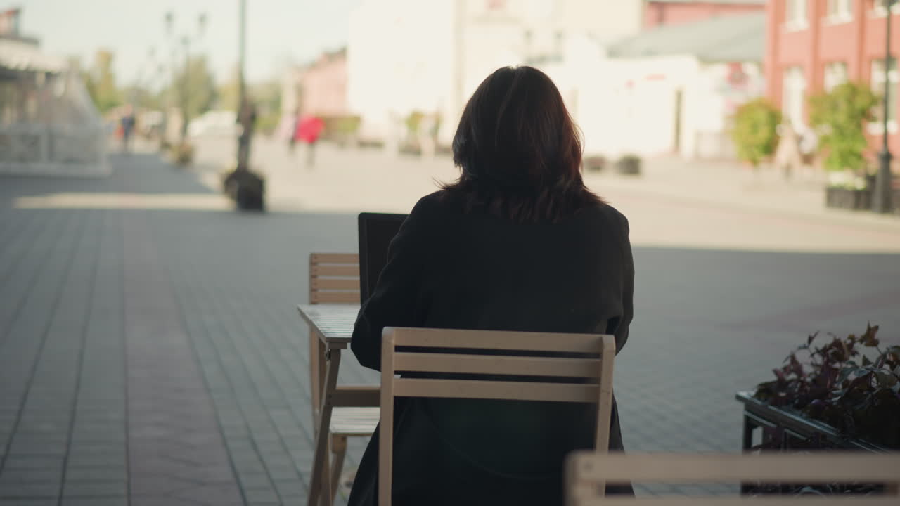 Rear view of woman working on laptop outdoors with her hair covering her face, decorative black pillar nearby, blurred urban background featuring modern buildings, and people walking in the distant