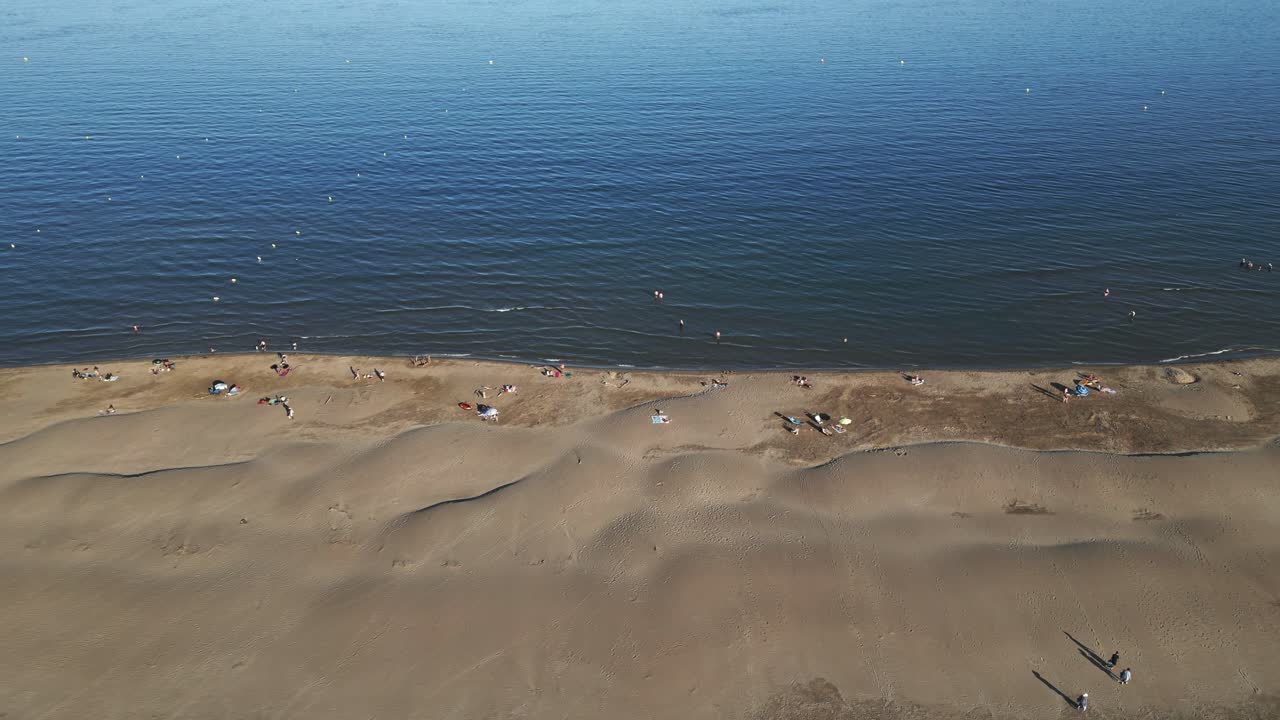 Families relax on a sandy shore under the sun, playing and swimming in the calm waters. Children build sandcastles, enjoying a perfect summer day. Aerial drone travelling