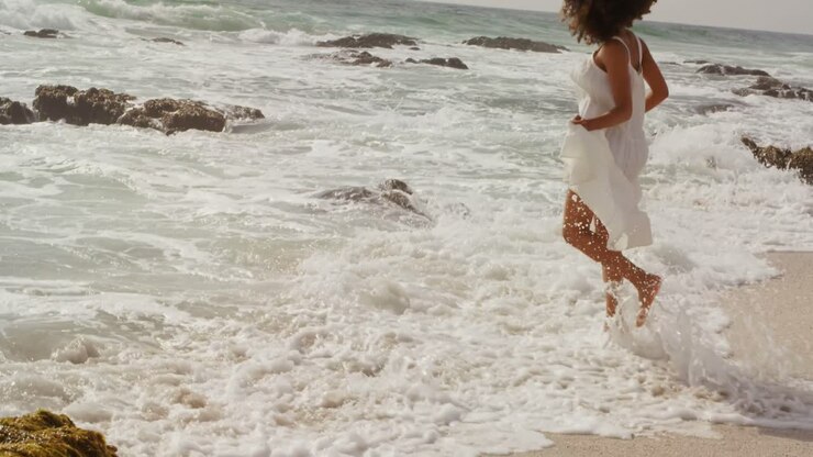 Vista laterale di donna afroamericana che gioca con l'acqua del mare sulla spiaggia 4k