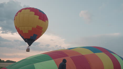 crew prepping second hot air balloon while earlier colorful balloon hovers above grassy open field at sunset during preflight setup capturing teamwork and scenic adventure mood under dramatic sky