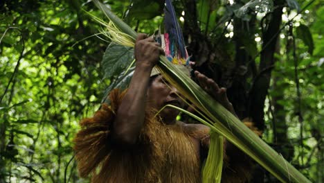 An-indigenous-man-wearing-a-feathered-hat-and-fringed-shirt-peels-young-coconut-leaves-in-Leticia,-Colombia