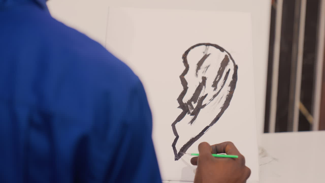Pittura nera su tela, vista da dietro di un artista che realizza tratti ampi e audaci con una matita verde, camicia blu, linee energiche che formano la silhouette di un orecchio, sfondo drammatico dello studio e illuminazione calda.