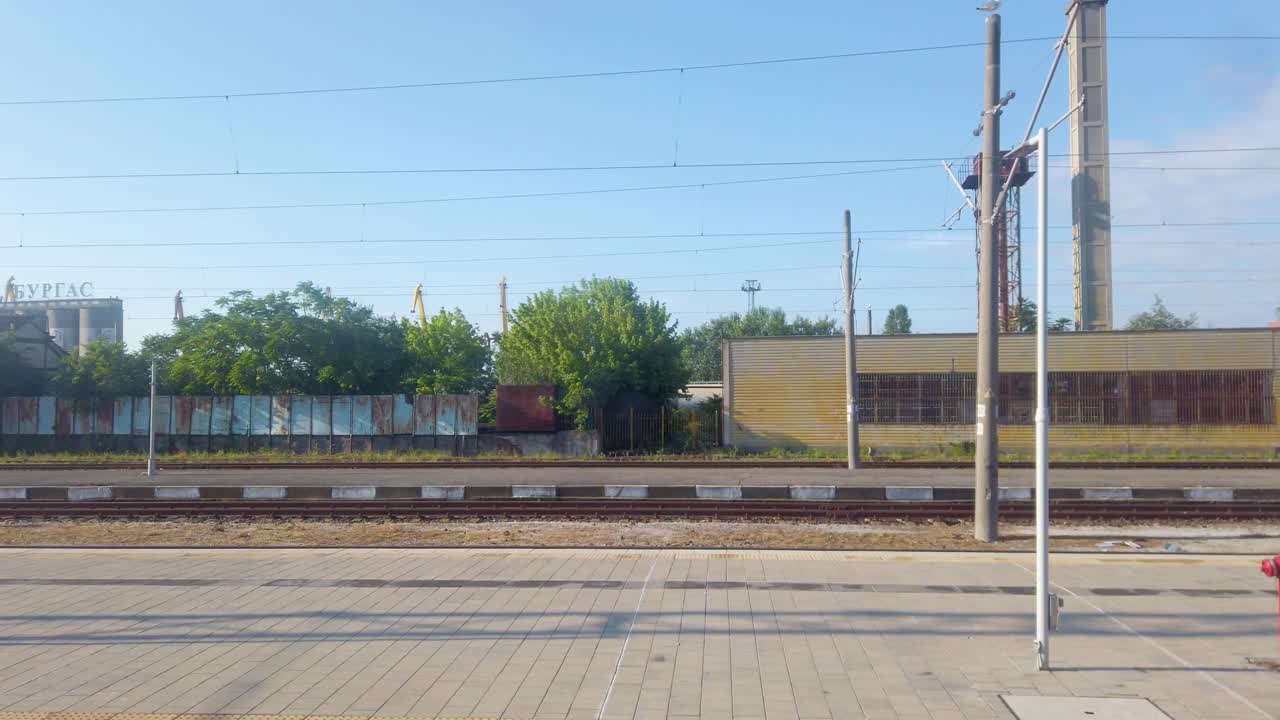 View of train station while the train is moving, traveling on the railroad in good sunny weather. BDZ. Rail transport in Bulgaria. View of trees and industrial buildings.