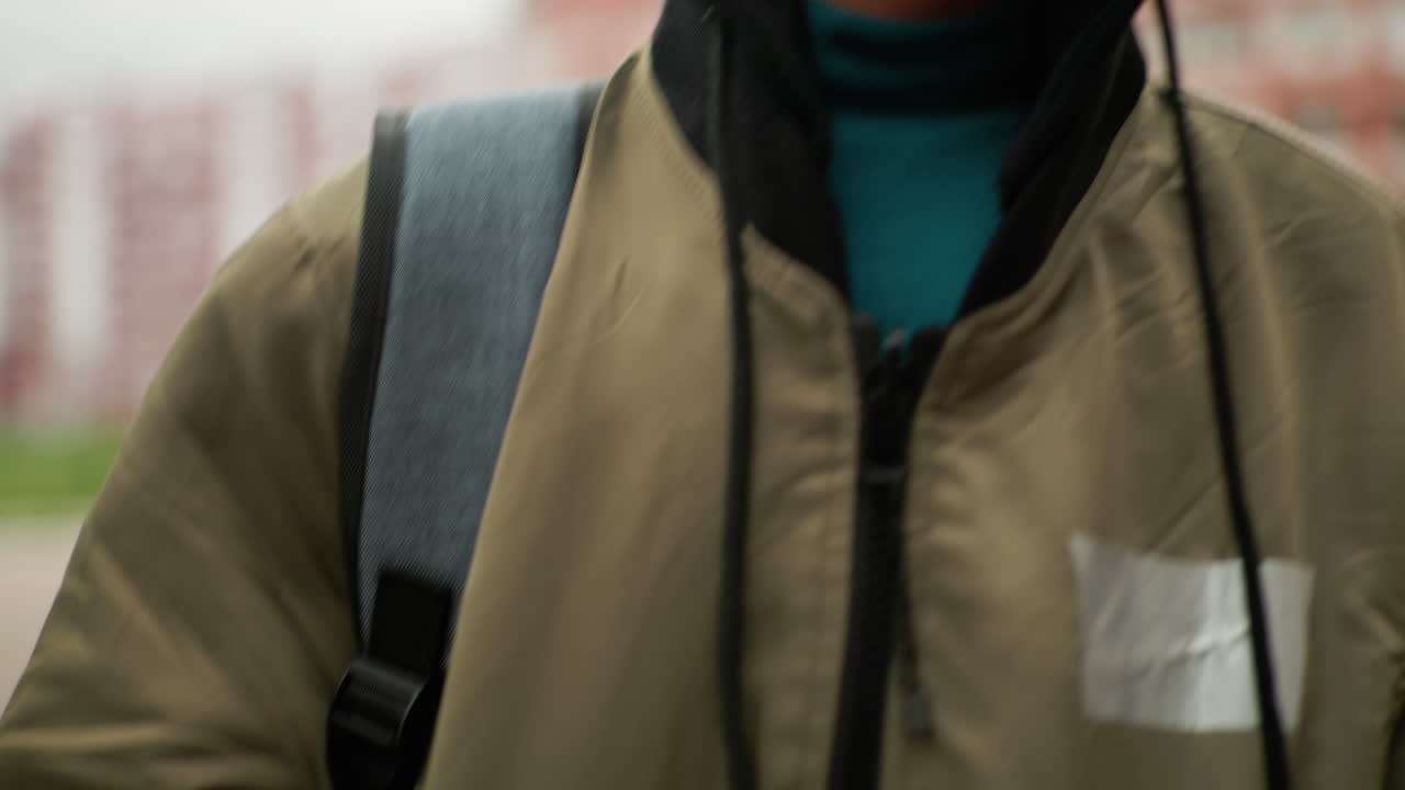 캠퍼스 구역에서 야외를 걸으며 가방끈을 조절하는 아이의 클로즈업, 일상적인 학교생활과 젊음이 느껴지는 학업 라이프스타일을 강조하는 흐릿한 주거용 건물 배경