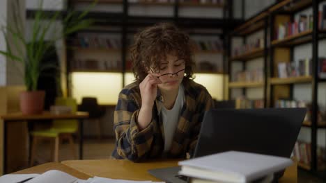 una estudiante cansada con el cabello rizado se quita las gafas durante sus estudios en la computadora portátil en la mesa de la biblioteca