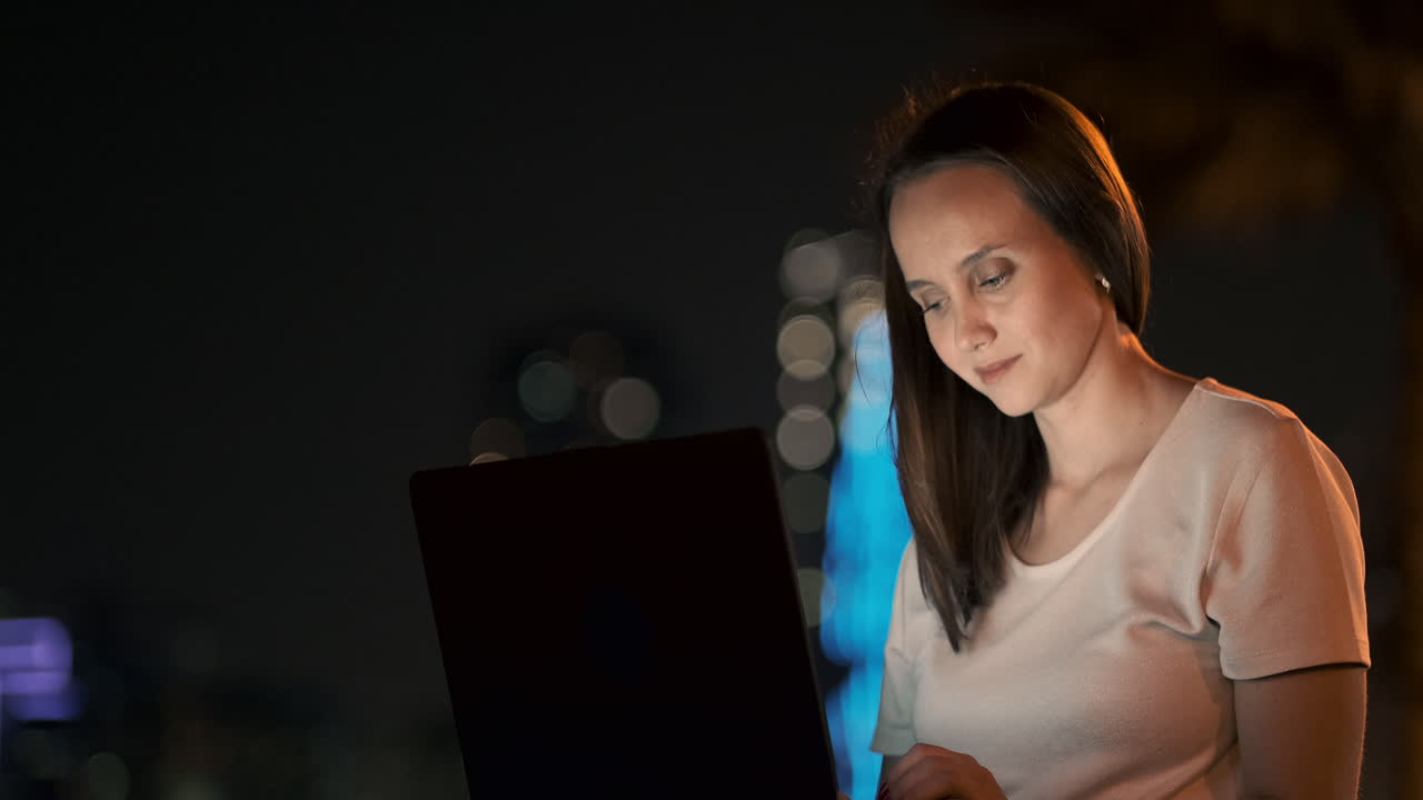 una joven estudiante con una computadora portátil por la noche en la ciudad mirando la pantalla de la computadora y escribiendo con las manos en el teclado. trabajo remoto en internet. la estudiante hace el trabajo. vida nocturna en la ciudad