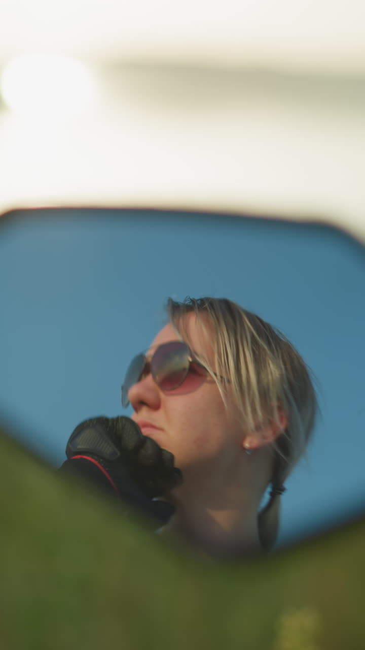 Mirror reflection of a woman wearing sunglasses, resting her chin on her hands while gazing intently at something, the background shows a soft blur of a field and water