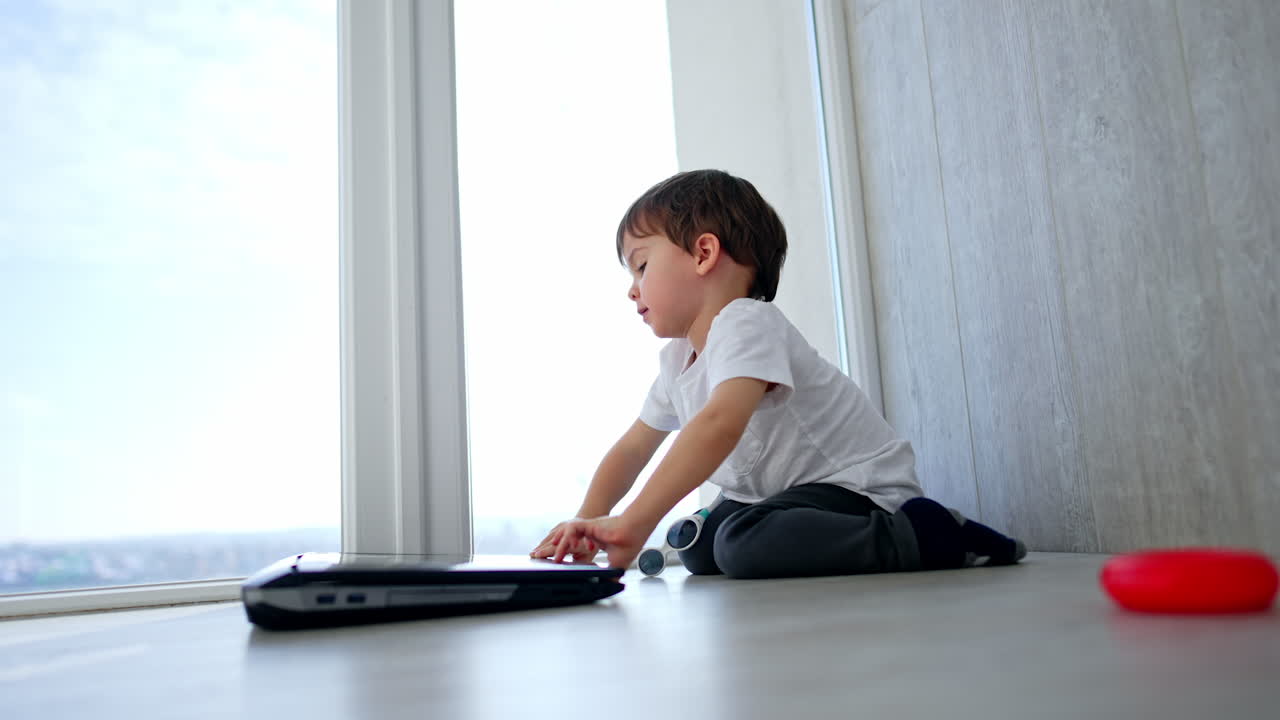 Beautiful Caucasian baby boy in white t-shirt and dark pants sits on the floor near laptop. Kid opens gadget smiling to camera. Low angle view.