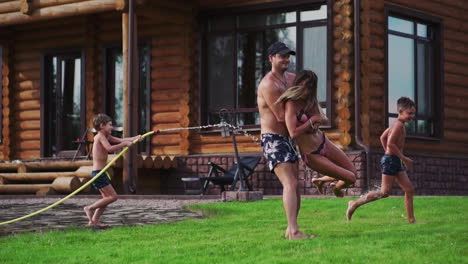 the two boys are playing with mom and dad in the backyard of their house drenching with hose water and laughing and smiling in slow motion. happy family