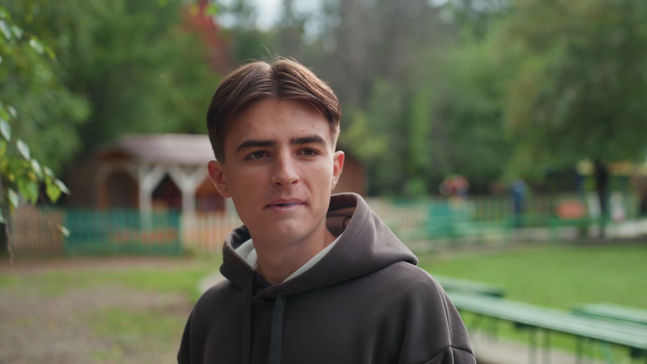 blanke tienerjongen met hoodie in park, beschouwend portret met zacht zonlicht, groene banken en gazebo op de achtergrond, openhartig vape-gebaar, wisselende uitdrukkingen van glimlach tot peinzend, filmisch