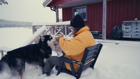 a person with his alaskan malamute dog seated outside in deep snow, pouring hot coffee on a mug