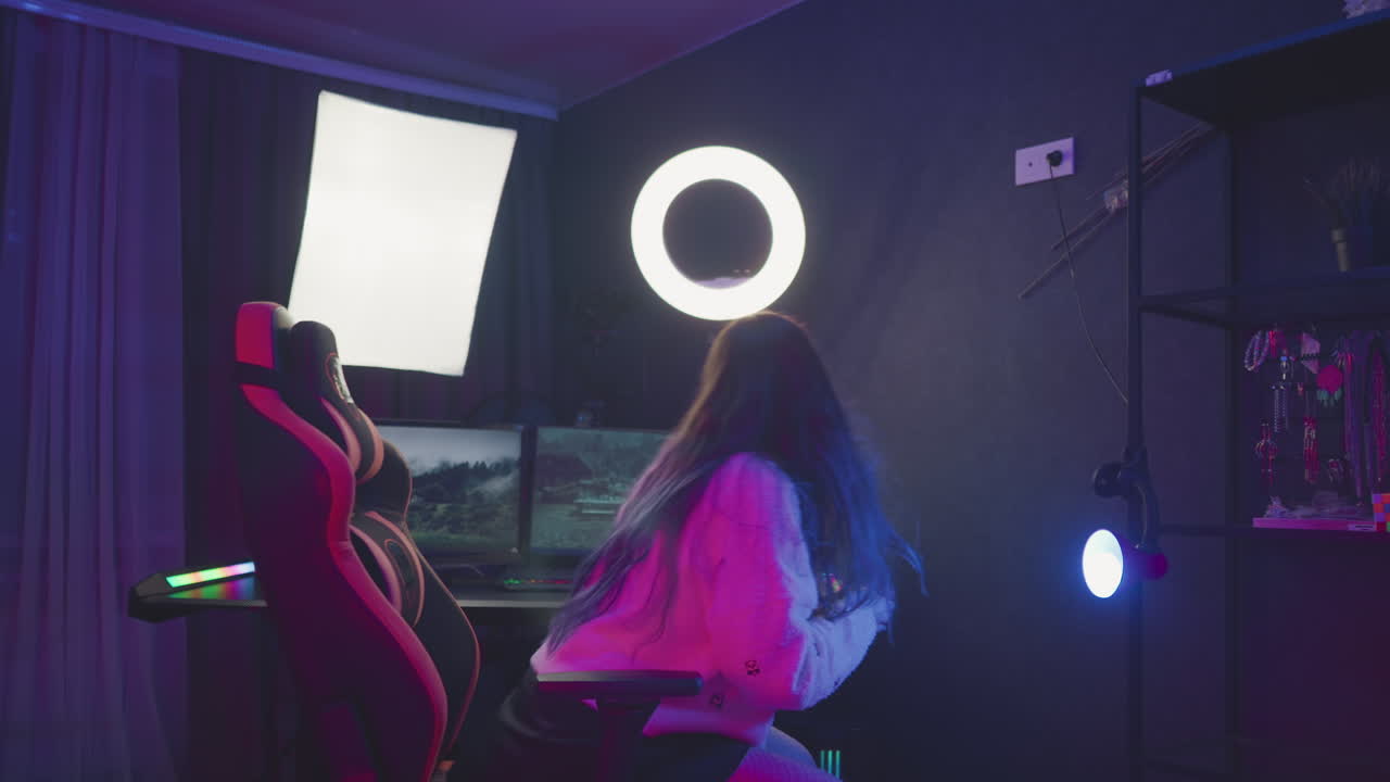 Woman walks toward ergonomic gaming chair in front of dual monitor setup with popcorn in hand, preparing to sit and adjust seat under soft ring and panel lighting in cozy tech studio environment