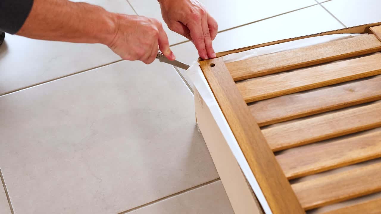 Hands carefully cut open a wooden slatted box on a tiled floor using a knife, highlighting precision and focus