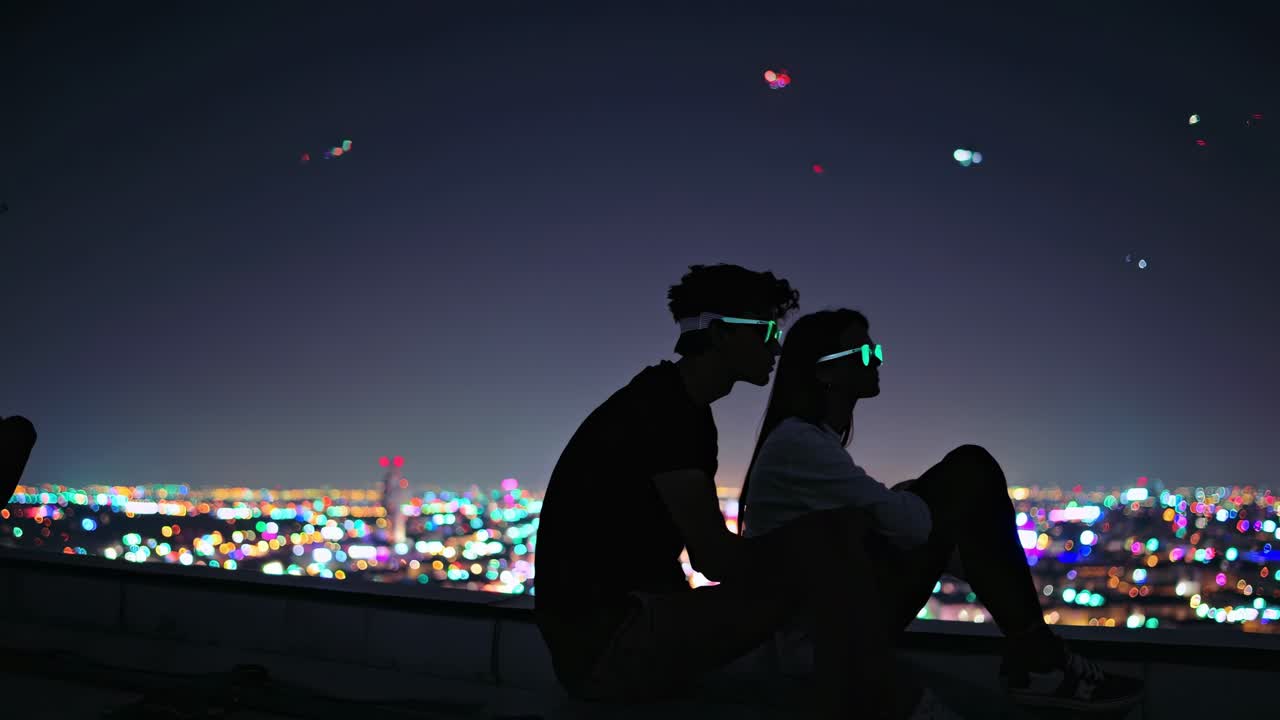 Stylish couple wearing illuminated glasses perched on urban rooftop, overlooking sparkling Los Angeles nightscape with vibrant aerial elements floating overhead