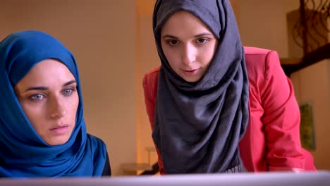 closeup portrait of two young arabian female employees arguing over the topic using the laptop indoors on the workplace