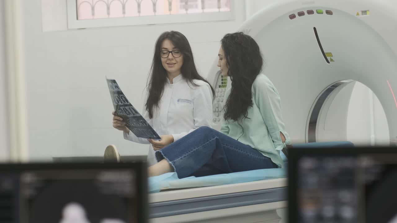 Woman doctor radiologist explains the results of CT scanning for young female patient, showing the snapshot with images, observing and analyzing CT scan in modern clinic beside modern CT scanner