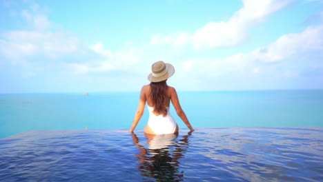 sitting on the edge of an infinity-edge pool a woman with her back to the camera raises her arms in joy to the ocean horizon