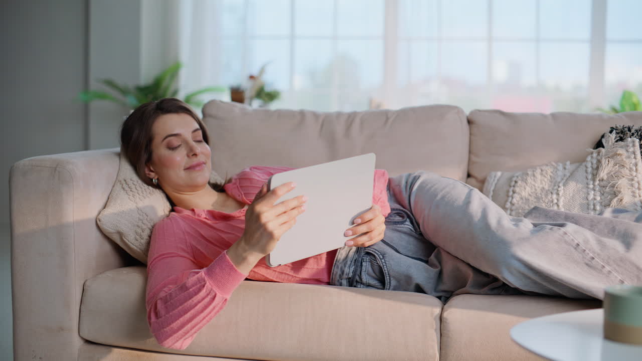 Primo piano di una donna calma che legge un ebook distesa sul divano accogliente di un appartamento al mattino