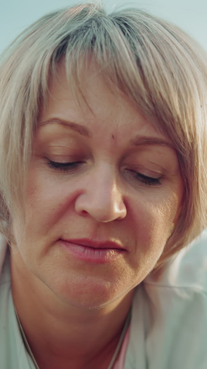 mujer serena de mediana edad con cabello gris acostada, piernas levantadas hacia atrás, ojos cerrados, apareciendo profundamente en pensamiento contra un fondo de campo abierto borroso, exudando paz y reflexión tranquila