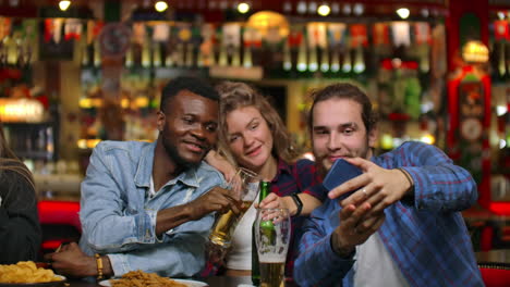 african american with european friends sitting in a bar takes a selfie. a large group of friends sits in a barz at one table chatting drinking beer and taking pictures.