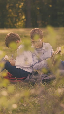 joyful schoolmates do home assignments after lessons sitting on fresh green lawn in spring sunny park