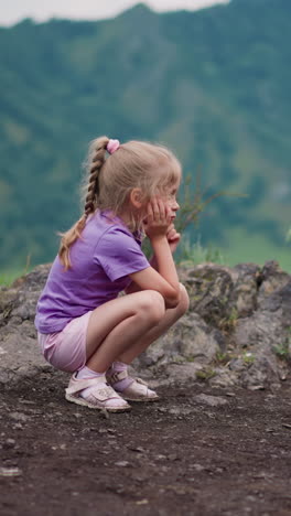 tired little girl sits on haunches leaning head on palm on rocky hill against distant mountains at national reserve on nasty day slow motion